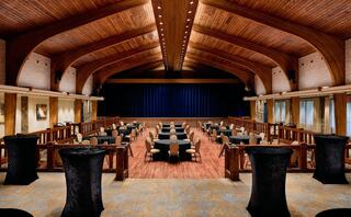 A spacious event hall featuring tall wooden ceilings, elegant round tables, and a stage with a blue curtain in a stylish and inviting setting.