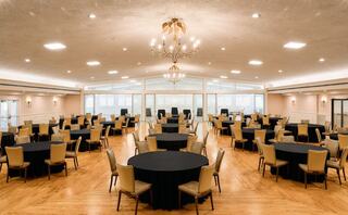 A spacious event hall features multiple round tables with black tablecloths, surrounded by beige chairs, illuminated by a chandelier.