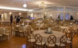 A spacious event venue features elegantly set tables with golden chairs, twinkling lights overhead, and a festive, inviting atmosphere.