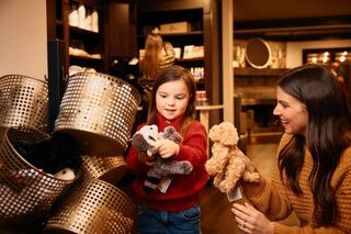 A young girl and a woman enjoy selecting stuffed animals from a basket in a cozy, stylish store, sharing smiles and laughter.