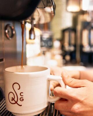 A barista pulls espresso into a white mug adorned with a logo, wearing a brown apron that reads "Coffee & Goods."