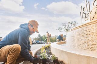 A person plants flowers near a fountain, focused on their task, while others work in the background under a partly cloudy sky.