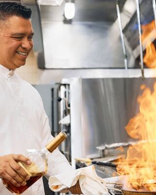 A chef smiles while pouring a liquid into a pan, creating a burst of flames in a busy kitchen environment.
