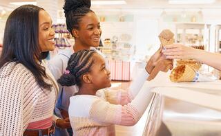 A joyful family of three smiles at an ice cream counter, eagerly waiting for a treat from a staff member. Bright and cheerful setting.