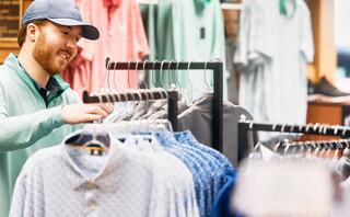A man smiles while browsing a rack of shirts in a retail store, surrounded by various clothing options.