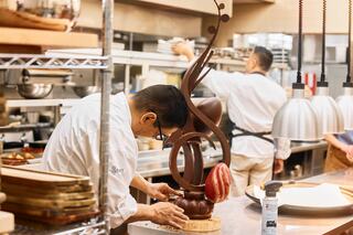 A person sprinkles fresh basil and cherry tomatoes onto a pizza base topped with tomato sauce and shredded chicken, preparing to bake.