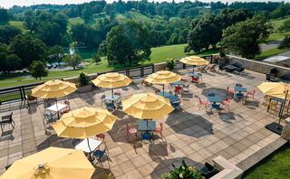 A spacious outdoor terrace features yellow umbrellas over tables, set against a lush green landscape and gentle hills in the background.