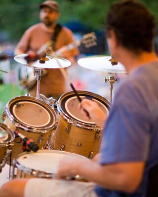 A drummer focuses on his kit while a guitarist plays in the background, capturing a lively musical moment outdoors.