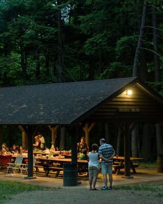 A couple stands close together outside a lit shelter, where a gathering and picnic are underway amidst a wooded area at dusk.