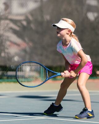 A focused young girl in a cap and colorful outfit prepares to play tennis, holding a racket on a sunny court.