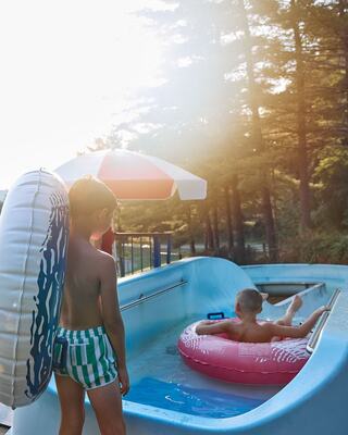 Two boys enjoy a sunny day at a water slide. One relaxes in a pink float while the other stands with a large inner tube by the water.