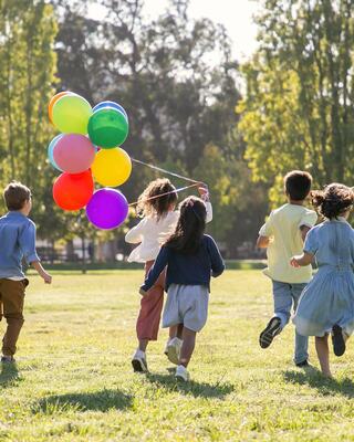 A group of children runs joyfully across a grassy field, one holding colorful balloons as sunlight filters through the trees.