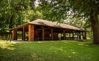 A spacious wooden pavilion nestled in a green park, surrounded by trees, with picnic tables visible under the shade for gatherings.