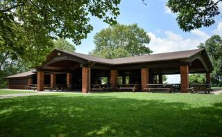 A spacious, rustic pavilion surrounded by green grass and trees, featuring picnic tables and a charming wood structure under a bright sky.