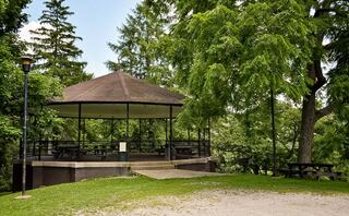 A shaded gazebo with a brown roof, surrounded by lush greenery and trees, offers a peaceful outdoor gathering space.