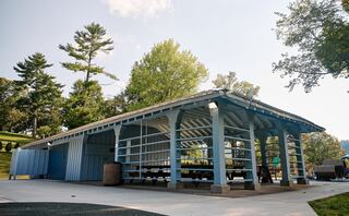 A blue pavilion with open sides sits amidst greenery, offering shaded seating near a playground. Trees provide a serene backdrop.