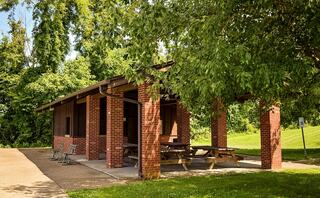 A brick shelter surrounded by lush greenery, featuring picnic tables and a concrete floor, perfect for gatherings and outdoor relaxation.
