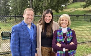 Three smiling individuals stand together outdoors, dressed warmly, surrounded by trees. A friendly and cheerful atmosphere exudes from them.
