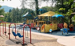 A vibrant playground features colorful structures, slides, and swings, surrounded by green trees and hills in a sunny outdoor setting.