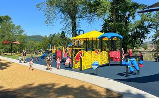 A vibrant playground filled with children and adults, featuring colorful equipment under bright sunshine, surrounded by trees and hills.