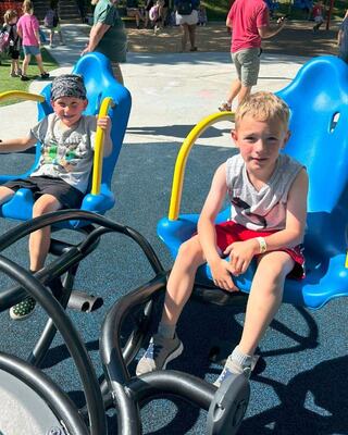 Two young boys sit on bright blue playground equipment, enjoying a sunny day. People can be seen in the background, creating a lively atmosphere.