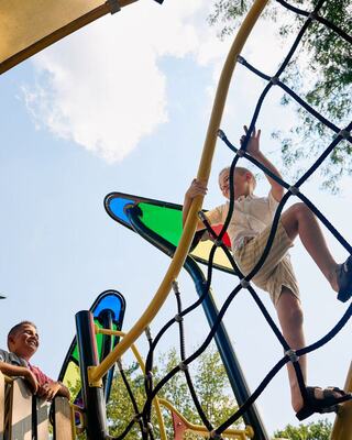Two children play on a colorful playground structure, one climbing while the other watches, under a bright blue sky with fluffy clouds.