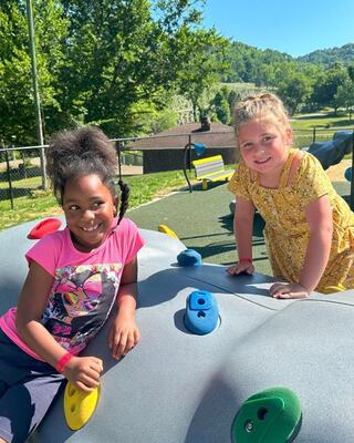 Two smiling children play on a colorful climbing structure at a sunny playground, surrounded by trees and rolling hills.
