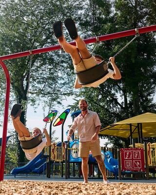 Two kids are swinging joyfully while a man watches and smiles, surrounded by vibrant playground equipment under a sunny sky.