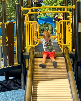 A young child in a shirt and red shorts joyfully slides down a playground slide, gripping the side for balance.
