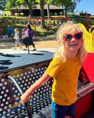 A smiling child in a yellow shirt and sunglasses stands on playground equipment, surrounded by other kids playing in a sunny park.