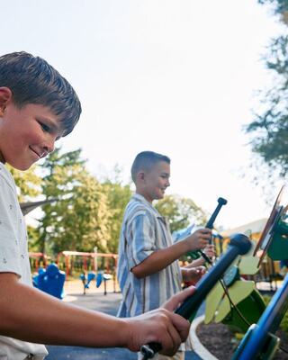 Two boys play joyfully on musical instruments at a sunny playground, surrounded by greenery and colorful equipment.