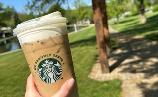 A hand holds an iced drink from Starbucks, set against a sunny park scene with a path and lush greenery.
