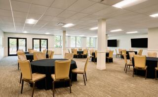 A spacious event room featuring round tables with black tablecloths, beige chairs, large windows, and modern decor.