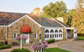 A charming stone building features a red awning, large arched windows, and vibrant flower arrangements, surrounded by lush greenery.