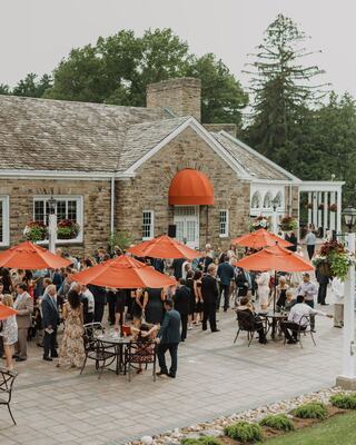 A lively outdoor gathering takes place at a charming venue, featuring guests mingling under orange umbrellas amidst lush greenery.