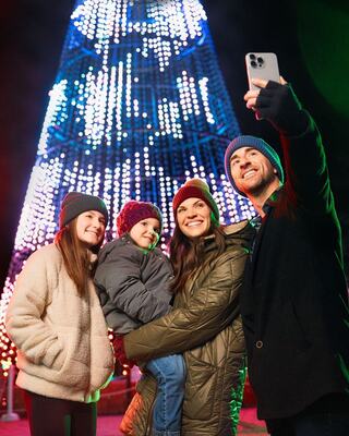A family joyfully takes a selfie in front of Oglebay's Welcome Tree at Festival of Lights, celebrating the holiday spirit together.