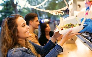A young woman happily receives a fresh salad from a food vendor, while two friends look on, enjoying the vibrant outdoor atmosphere.