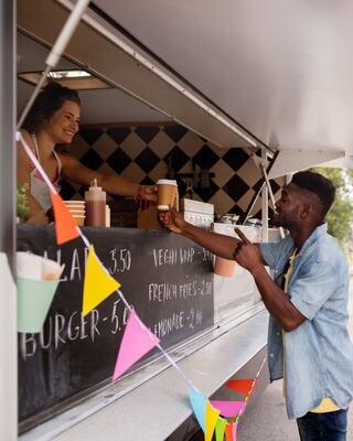 A smiling vendor hands a cup to a customer at a food truck, with colorful decorations and a menu visible in the background.