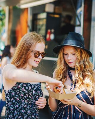 Two young women share food from a cardboard container, enjoying a sunny day at a food market. One wears sunglasses, the other a hat.