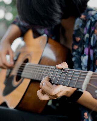 A person playing an acoustic guitar, focusing intently on their fingers as they strum, wearing a floral-patterned shirt.