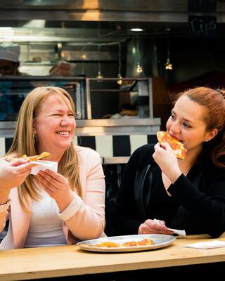 Two women enjoy pizza at a casual eatery, sharing laughter and food, with a chef visible in the background, creating a lively atmosphere.