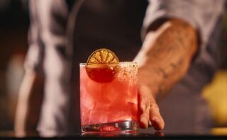 A vibrant pink cocktail garnished with a dried lime slice, held by a bartender with a tattooed arm, exuding a lively bar atmosphere.