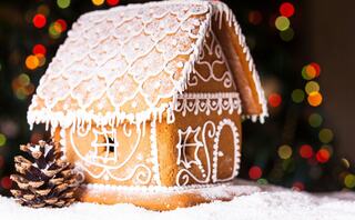 A beautifully decorated gingerbread house with intricate icing details, surrounded by powdered sugar and a pinecone, set against festive lights.