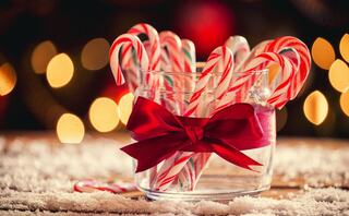 Colorful candy canes in a glass jar, tied with a red ribbon, set against a blurred, festive background of warm holiday lights.
