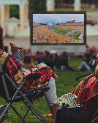 Two people are seated on a lawn, wrapped in blankets, enjoying a large outdoor screen showing a crowded event.