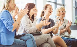 A group of women sits together, smiling and clapping, clearly engaged and enjoying the moment. The atmosphere feels positive and supportive.