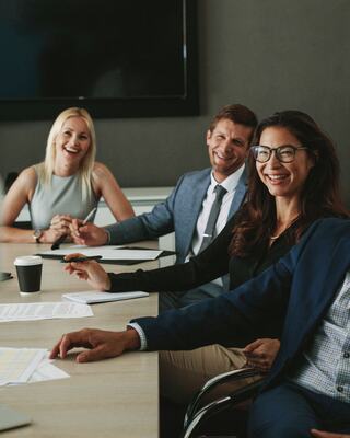 A group of diverse individuals are engaged in a moment of applause, with a woman in a blazer prominently smiling in appreciation.