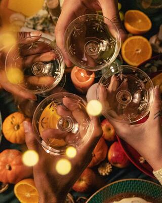 Four hands hold elegant glasses, surrounded by vibrant fruits and soft, warm lighting, creating a festive and cheerful atmosphere.