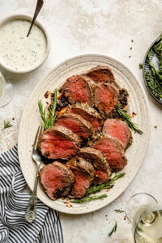 Slices of perfectly cooked beef are arranged on a platter, garnished with herbs and accompanied by a creamy sauce and wine.