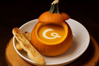 A pumpkin hollowed out to serve creamy soup, topped with a whimsical design, accompanied by toasted breadsticks on a plate.